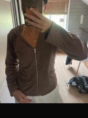 Brun zip hoodie med huva - Säljer en brun hoodie med dragkedja framtill och huva. Hoodien har en smal passform och är gjord i ett mjukt, sammetsliknande material. Perfekt för dig som gillar en enkel men snygg stil.