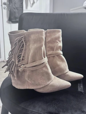 Beigea bootsmed fransar - Säljer ett par beigea boots i mocka med spetsig tå och dekorativa fransar på sidan. Skorna har en låg klack och ett band runt ankeln för extra detalj. Dragkedja på insidan och klacken är 10cm, har köps för 600 har använts bara en gång💕