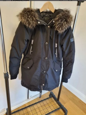 Svart Rock and blue dunjacka   - Snygg svart parkas med stor huva och fluffig. Jackan har dragkedjor och knappar framtill, två bröstfickor med dragkedja och två snedställda fickor med lock. Perfekt för kalla dagar och riktigt varm. Lång modell som går ner över låren. Väldigt lite använd. 