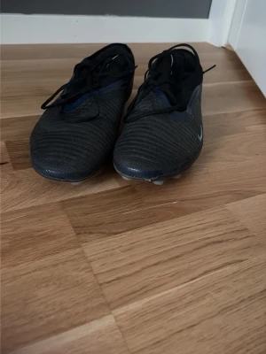 Svarta fotbollsskor Nike elit - Säljer ett par svarta fotbollsskor med snörning och låg profil. Skorna har en diskret design med små detaljer i mörkblått och en liten vit logga på sidan. Ovandelen är i syntetmaterial med struktur för bättre bollkontroll. Perfekta för dig som vill ha stilrena skor på planen.