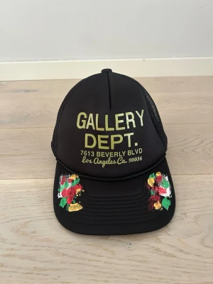 Svart truckerkeps Gallery Dept. - Snygg svart truckerkeps från Gallery Dept. med mesh baktill och justerbar snapback. Framtill står det 'GALLERY DEPT.' i gult tryck och kepsen har färgglada handmålade detaljer på skärmen. Perfekt för en streetwear-look.