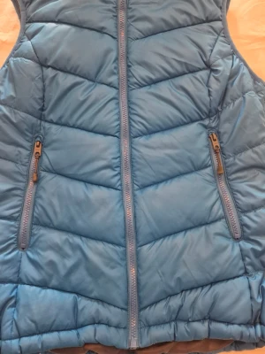 Blå dunväst med huva - Snygg blå dunväst med dragkedja framtill och två fickor med dragkedja. Västen har en stor, fodrad huva med dragsko och quiltad design. Perfekt för lager på lager och håller dig varm under kyliga dagar.