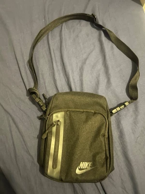 Svart axelväska från Nike - Snygg svart axelväska från Nike med justerbar rem och dragkedja framtill. Väskan har en stilren design med Nike-logga på framsidan och praktiskt ytterfack. Perfekt för att bära det viktigaste när du är på språng.