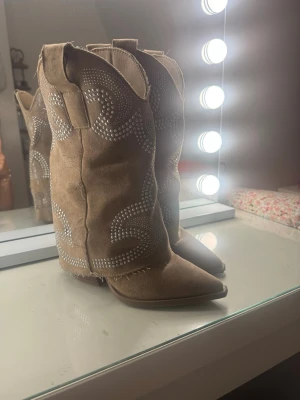 Beiga cowboy boots med nitar - Snygga beiga cowboy boots i mockaimitation med spetsig tå och bred klack. Dekorerade med silvriga nitar i mönster och har råa kanter upptill. Dragkedja på insidan för enkel påtagning. Perfekta för en cool westernstil.