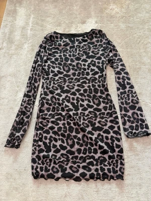 Leopardmönstrad långärmad klänning - Snygg långärmad klänning med leopardmönster i svart och beige. Klänningen har en kroppsnära passform och är tillverkad i ett stretchigt, tunt material. Rund halsringning och vågig kant nertill ger en trendig look.