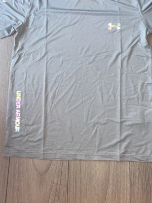 Grå Under Armour t-shirt - Snygg grå t-shirt från Under Armour med färgglad logga på bröstet och text längs sidan. Tillverkad i ett lätt och funktionellt material som passar perfekt till träning. Klassisk passform och korta ärmar.