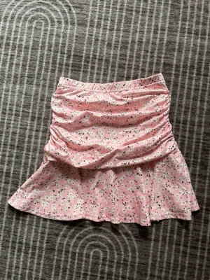Rosa blommig kortkjol med volang, 134 - Söt rosa kortkjol med blommigt mönster och volangkant. Kjolen har rynkade detaljer upptill och är gjord i ett mjukt, lätt material som passar perfekt till varma dagar. Elastisk midja för skön passform.