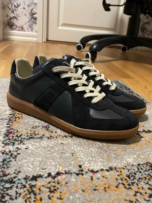 Maison Margiela Gats - Snygga svarta sneakers med ovandel i både läder och mocka, samt kontrasterande vita snören. Skorna har en klassisk låg profil och en brun gummisula som ger en retrovibe. Perfekta för dig som gillar stilrena och tidlösa sneakers.