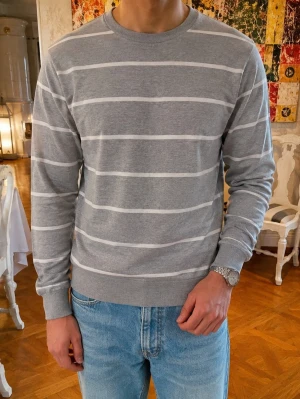 Vintage Kashmir Grå randig tröja från Paul Rosen - Snygg grå tröja från Paul Rosen med vita horisontella ränder. Tillverkad i en mjuk mix av pima cotton och cashmere för extra komfort. Rund halsringning och ribbade muddar vid ärmslut och nederkant. Perfekt till jeans eller chinos.