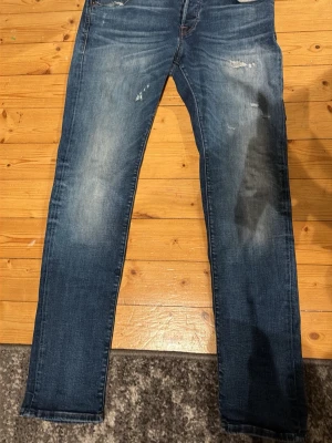 Glenn slim fit jeans blå slitningar - Säljer ett par blå slim fit jeans med slitningar och tvättade detaljer på låren och knäna. Jeansen har klassisk femficksdesign och normal midja. Perfekta för en avslappnad och trendig look.