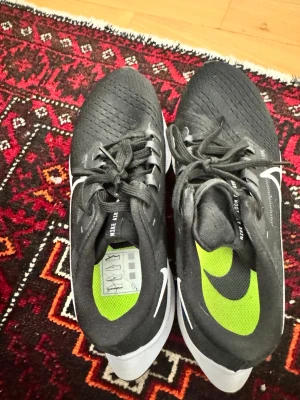 Svarta Nike sneakers med vit sula - Snygga svarta Nike sneakers med vit sula och grönt innerfoder. Skorna har klassisk låg profil, ventilerande mesh-ovandel och vita detaljer på sidan. Perfekta för dig som gillar sportig och stilren look.