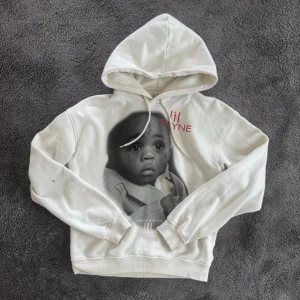 Vit hoodie med Lil Wayne-print - Säljer en vit hoodie med stor huva och dragsnören. På framsidan finns ett stort tryck av Lil Wayne som barn samt texten 'Lil Wayne' i rött. Hoodien har ribbade muddar vid ärmar och midja. Perfekt för dig som gillar streetwear och hiphop. Storlek xs egentligen men passar mig som bär storlek M (183cm, 67kg)