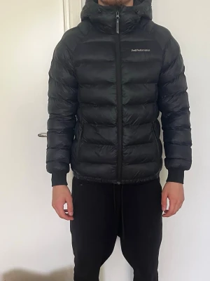 Svart dunjacka från Peak Performance - Säljer en svart dunjacka från Peak Performance med huva och dragkedja framtill. Jackan har en puffig design med breda kanaler, två sidofickor med dragkedja och diskret logga på bröstet. Perfekt för kalla vinterdagar.
