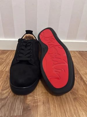 Louboutin skor  - Stilrena svarta sneakers med klassisk låg profil och snygg röd sula. Skorna har svarta skosnören, ovandel i mockaliknande material och ljus insida. Perfekta för dig som vill ha en clean look med en twist.