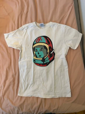 Vit Billionaire Boys Club t-shirt astronaut - Vit t-shirt från Billionaire Boys Club med stort tryck av en astronaut på framsidan i rött, turkost och svart. Klassisk passform och rund halsringning. Tillverkad i 100% bomull, perfekt för en streetwear-look.