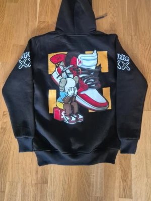 Svart  hoodie med tryck - Svart hoodie med stor färgglad grafisk print på ryggen som visar en tecknad figur och sneakers. Tryck med 'Off-White' och 'XX' på båda ärmarna samt bröstet. Klassisk känguruficka och huva med dragsko. Perfekt för streetwear-stilen. Strl s men mer som M