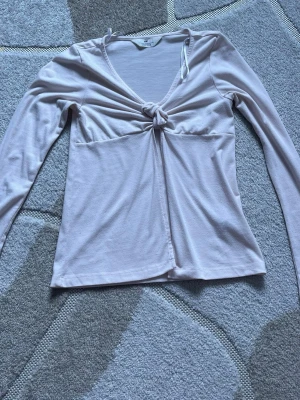 Ljusrosa långärmad topp med knyt - Supersöt ljusrosa långärmad topp med v-ringning och knyt framtill. Toppen har en mjuk och stretchig känsla, perfekt för att styla med jeans eller kjol. Enkel men ändå unik detalj med knuten framtill som ger en trendig look.