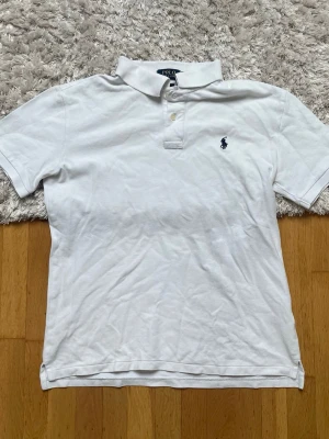 Vit pikétröja från Polo Ralph Lauren - Klassisk vit pikétröja från Polo Ralph Lauren med broderad logga på bröstet. Tröjan har korta ärmar, krage och knappar framtill. Modellen är custom fit och passar perfekt till en avslappnad men stilren look.