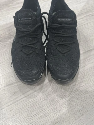 Nike Air Vapormax Flyknit 2023 - Svarta Nike Air Vapormax sneakers med stickad ovandel och synlig sula i plast. Skorna har en diskret grå Swoosh på sidan och snörning framtill. Lätta och flexibla med modern design, perfekta för dig som gillar streetwear och sportig stil.