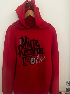Röd hoodie från Angelo Litrico - Snygg röd hoodie från Angelo Litrico med trycket 'Mitre Records Co Inc' i svart och vitt på bröstet. Hoodien har stor huva och magficka framtill. Tillverkad i mjukt material som passar perfekt för en avslappnad stil.