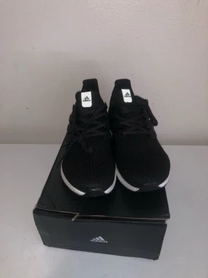 Svarta Adidas Ultraboost sneakers - Snygga svarta Adidas Ultraboost sneakers med stickad ovandel och vita Boost-sulor. Skorna har klassiska tre ränder på sidan, svart snörning och diskret Adidas-logga på plösen. Perfekta för dig som gillar sportig och stilren look.