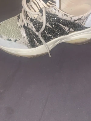 Steve madden skor  - Unika sneakers med glitterdetaljer, ormskinnsmönster och zebrarandig plös. Skon har vita och svarta inslag samt beige partier. Tjock sula och snörning framtill. Perfekt för dig som vill sticka ut med din stil.