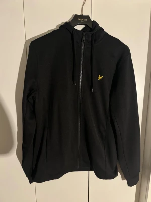 Svart hoodie från Lyle & Scott - Säljer en svart hoodie från Lyle & Scott med dragkedja framtill och justerbar huva. Klassisk gul logga på bröstet. Tillverkad i mjukt material, perfekt för en avslappnad stil. Långa ärmar och fickor på sidorna.