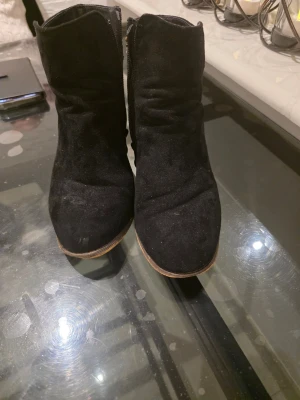 Svarta stövletter med träklack - Snygga svarta stövletter i mockaimitation med grov träklack och guldfärgade detaljer baktill. Dragkedja på sidan för enkel påtagning och rundad tå. Perfekta för dig som vill ha en cool och trendig look.