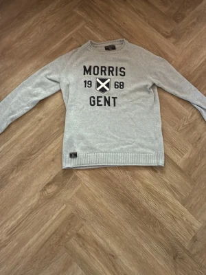 Ljusgrå tröja Morris Gent - Ljusgrå tröja från Morris med tryck 'Morris Gent 1968' och ett broderat emblem på bröstet. Tröjan har rund halsringning, långa ärmar och ribbade muddar vid ärmslut och nederkant. Perfekt för en avslappnad och stilren look.