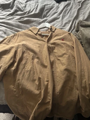 Beige skjorta från Burberry London - Klassisk beige skjorta från Burberry London med diskret röd logga broderad på bröstet. Skjortan har knappar framtill och en stilren krage. Tillverkad i mjuk bomull, perfekt för en clean och tidlös look. Passar large också, baggy passform 
