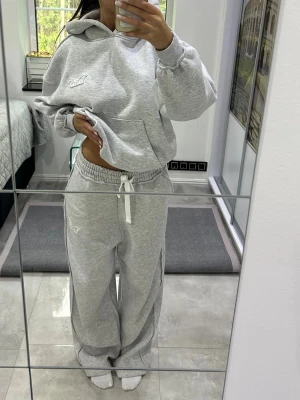 Grått loungewear-set med huva - Mysigt loungewear-set i ljusgrått med hoodie och matchande byxor. Hoodien har magficka, huva och broderad textdetalj på bröstet. Byxorna har resår i midjan med vit dragsko och broderad text på benet. Perfekt för chill dagar hemma.