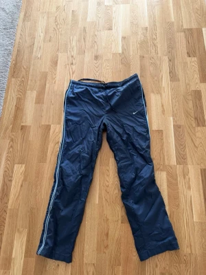 Nike byxor vintage - Passar bäst L. Snygga marinblå träningsbyxor från Nike med vita detaljer längs sidorna och dragkedja vid benslut. Byxorna har resår i midjan och är tillverkade i ett lätt, sportigt material som passar perfekt till träning eller uppvärmning.