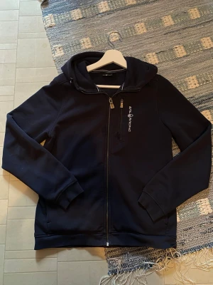 Marinblå hoodie från Sail Racing - Snygg marinblå hoodie från Sail Racing med dragkedja framtill och huva. Tröjan har ett broderat Sail Racing-tryck på bröstet och mjukt fleecefoder på insidan. Perfekt för dig som gillar sportig och stilren look.