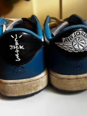 Nike Air Jordan 1 Low Travis Scott - Snygga Nike Air Jordan 1 Low Travis Scott x Fragment sneakers med svart, vit och blå färgskala. Skorna har ikoniska detaljer som omvänd swoosh, broderade loggor på hälen och blå insida. Materialet är en mix av läder och textil för en skön och stilren look.