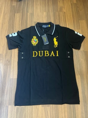 Polo Ralph Lauren - Dubai - Svart pikétröja från Polo Ralph Lauren med gul brodyr, stor gul logga och texten DUBAI framtill. Klassisk krage med vit kant och knappar. Detaljer på ärmarna och broderade emblem på bröstet ger en exklusiv känsla. Helt ny slim fit