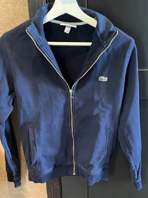 Marinblå ziptröja från Lacoste - Snygg marinblå ziptröja från Lacoste med klassisk krokodillogga på bröstet. Tröjan har hög krage, dragkedja framtill och två fickor. Tillverkad i mjukt bomullsmaterial och passar perfekt för en avslappnad stil.