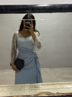 Ljusblå balklänning med glitterdetaljer - Elegant ljusblå långklänning med glittriga detaljer på överdelen och transparenta, glittriga långärmar. Klänningen har draperad midja och figurnära passform. Perfekt för speciella tillfällen när du vill sticka ut. Klänningen har använts en gång på ett bröllop och behövs inte längre. Den är skräddarsydd efter mina mått, men jag skulle säga att den är någonstans mellan storlek 36-38. Originalpriset på denna är 7000kr, säljs nu för 3500