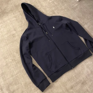 Mörkblå hoodie från Polo Ralph Lauren - Snygg mörkblå hoodie från Polo Ralph Lauren med dragkedja, huva med snörning och klassisk logga på bröstet. Bara använt några gånger så den är i bra skick 