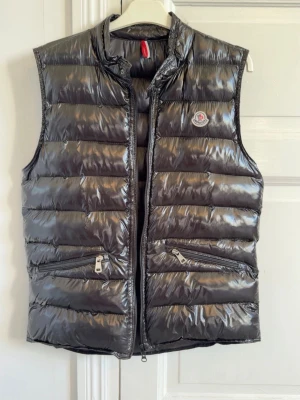 Svart Moncler väst - Snygg svart dunväst från Moncler med glansig finish och ståkrage. Västen har två dragkedjeförsedda fickor framtill och Moncler-logga på bröstet. Perfekt för lager-på-lager och håller dig varm med sin vadderade design.