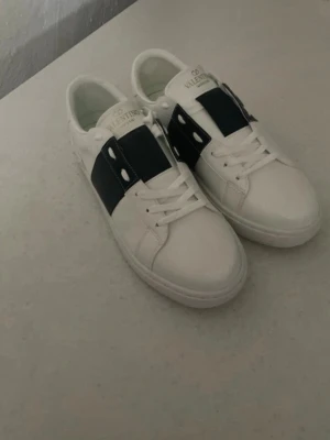 Vita sneakers med blå detalj - Snygga vita sneakers med bred blå rem över snörningen och vita nitar på sidorna. Klassisk låg modell med rund tå och vit sula. Perfekta för en trendig och stilren look.