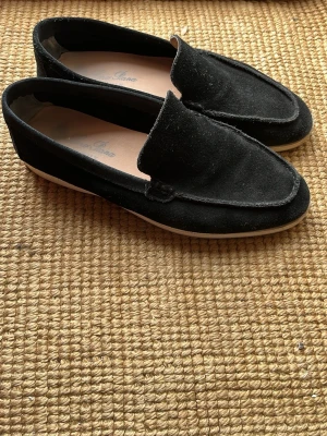 Loro Piana loafers - Säljer ett par stilrena svarta loafers i mocka med vit sula. Skorna har en klassisk design med rundad tå och diskret söm längs kanten. Perfekta för dig som vill ha en enkel men snygg sko till vardagslooken.