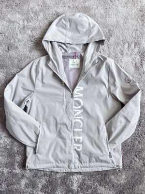 Ljusgrå vindjacka från Moncler - Snygg ljusgrå vindjacka från Moncler med luva och 2 fickor framme och en inuti jackan. Jackan har vit Moncler-logga längs dragkedjan och en patch på ärmen. Jackan är perfekt för hösten och den är vattentät. 