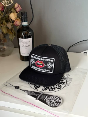 Svart Chrome Hearts Keps - Snygg svart keps från Chrome Hearts med en stor broderad patch framtill som visar röda läppar och vita detaljer. Kepsen har mesh på sidorna och justerbar snapback. Perfekt för dig som vill sticka ut med en unik accessoar.