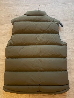 Olivgrön dunväst med hög krage - Snygg olivgrön dunväst med hög krage och tryckknappar framtill. Västen har två stora snedställda fickor och en liten broderad detalj på bröstet. Perfekt för lager på lager och riktigt varm tack vare den tjocka vadderingen.
