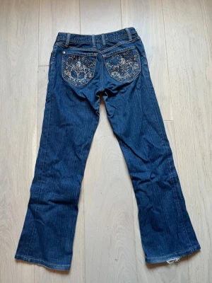 Y2k jeans med coola fickor - Snygga mörkblåa raka jeans med dekorativa bakfickor. Från märket Born In California. Storleken på lappen stämmer inte, utan de är omsydda för att passa barnstorleken 152/158 cm. Lite slitet längst ner och vissa av paljetterna har fallit av, men det är inget som jag tycker att man tänker på