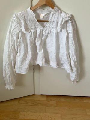 Vit blus med volanger från Gina Tricot - Söt vit blus från Gina Tricot med volangdetaljer och spetsinslag. Blusen har v-ringning, långa ärmar med resår vid ärmslut och en luftig passform. Perfekt för en trendig och romantisk look.