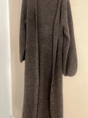 Lång brun stickad kofta - Mysig lång kofta i brunmelerat stickat material. Koftan har knappar framtill, långa ärmar och en rak, avslappnad passform. Perfekt att slänga över en outfit för extra värme och stil.