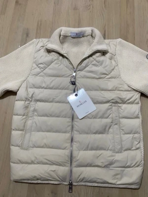 Krämvit dunväst från Moncler - Snygg krämvit dunväst från Moncler med stickade partier på ärmar och rygg. Västen har dragkedja framtill, hög krage och två fickor med dragkedja. Perfekt för lager-på-lager och ger en exklusiv känsla till din outfit.