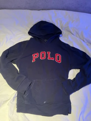 Polo Ralph lauren hoodie - Mörkblå hoodie med stor röd POLO-text på bröstet. Klassisk modell med huva och magficka. Mjukt material och ribbade muddar vid ärmslut och nederkant. Perfekt för en avslappnad och sportig stil.