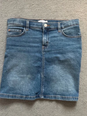 Blå jeanskjol från Name It - Säljer en klassisk blå jeanskjol från Name It i storlek 158. Kjolen har fyra fickor, kjolen stängs med knapp och dragkedja. Har aldrig använt den.🩷